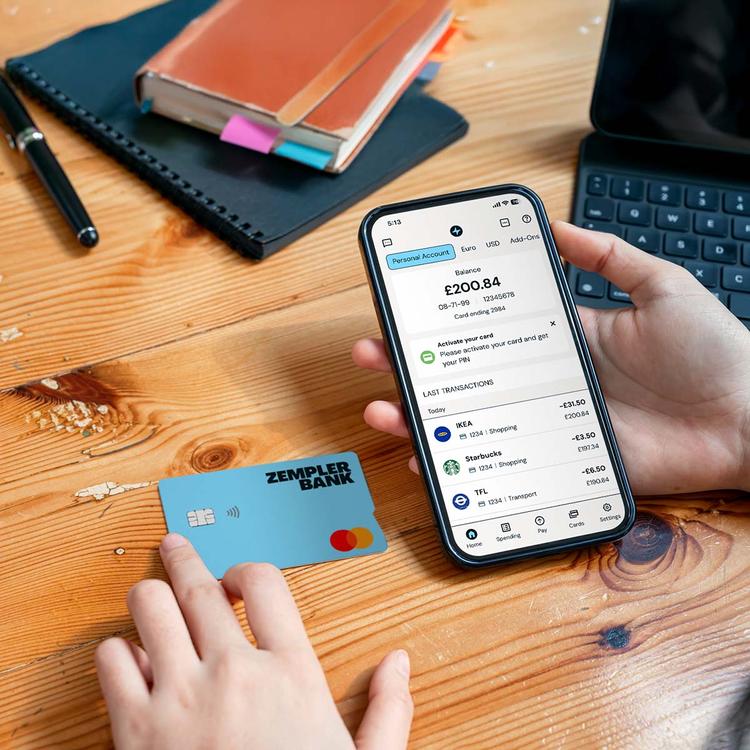 One hand holding a mobile phone showing the Zempler Bank app beside the Zempler Bank card